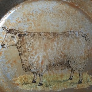 May include: A decorative plate with a sheep illustration. The plate has a metallic gold finish with a distressed look. The sheep is depicted in a field of grass, with brown and tan tones.