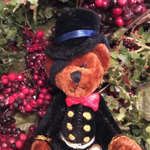 May include: A plush teddy bear ornament, dressed in a black suit with gold buttons and a red bow tie. The bear wears a black top hat with a blue band. The ornament is surrounded by greenery and red berries.
