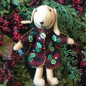 May include: A handmade plush bunny doll with long ears, wearing a plaid coat decorated with holly leaves and berries. The coat is buttoned with a single, round button. The doll is surrounded by greenery and red berries.