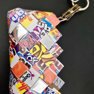 May include: A woven wallet made from colorful candy wrappers. The wallet features a variety of brand logos and designs, with a silver-colored clasp and a brown leather strap with a silver key ring.