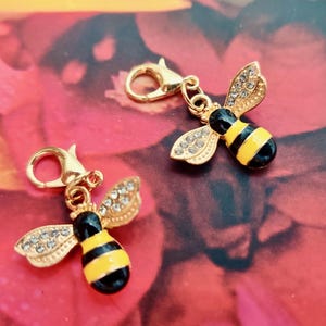 May include: Two gold-colored bee charms with lobster clasps. The bees have black and yellow striped bodies, gold wings with clear crystal accents, and black heads. The charms are set against a blurred floral background.