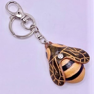 May include: A leather bee keychain with a silver clasp and key ring. The bee has a yellow body with black stripes and brown wings with a detailed pattern. A small silver stud is on the bee's head.