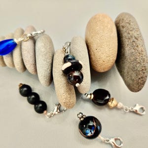 May include: An assortment of handmade jewelry charms with silver clasps. The charms feature various dark-colored beads, including black, brown, and blue, and are displayed with smooth, gray stones. The charms are designed for jewelry making.
