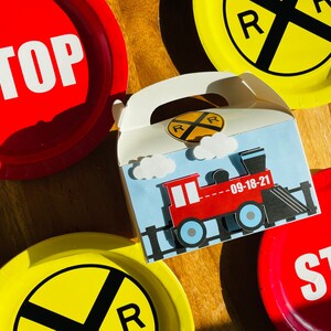 May include: A train-themed gift box with a red train illustration, clouds, and the date "09-18-21" on a light blue background. Red and yellow paper plates with "STOP" and railroad crossing symbols surround the box.