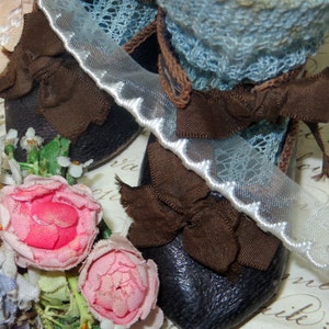 May include: Close-up of miniature black leather shoes with brown bows, paired with a blue lace trim and white scalloped ribbon. Pink fabric roses and other floral accents are scattered around the shoes, creating a vintage aesthetic.
