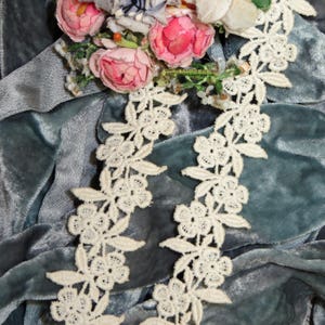 May include: White lace trim with a floral design on a blue velvet background. The lace trim is made of delicate, intricate flowers and leaves.