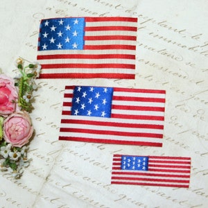 May include: Three miniature American flags made of fabric with red, white, and blue stripes and blue stars. The flags are arranged in a vertical stack, with the largest flag at the top and the smallest flag at the bottom.