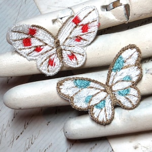 May include: Two embroidered butterfly patches. One has white wings with red dots, the other has white wings with blue dots. Both have brown thread outlining the wings and body. The patches are on a white surface.