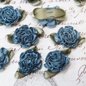 May include: Close-up of several teal blue fabric rose embellishments with olive green satin ribbon leaves. The roses have a ruffled texture and are arranged on a vintage paper background. These decorative floral accents are ideal for craft projects.