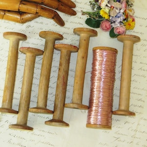 May include: A collection of vintage wooden spools and a spool of copper wire. The wooden spools have a mushroom-like shape with a wide base and a narrow top. The copper wire is tightly wound around a wooden core. A wooden articulated hand and flowers are in the background.