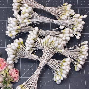 May include: Bundles of white and yellow tipped stamens, tied together with brown wire. The stamens are arranged on a black surface with a grid pattern. A few pink and green floral accents are visible in the lower left corner.