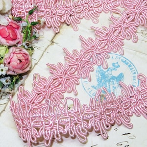 May include: Close-up of a delicate pink lace trim with a looped design. The trim is made of a soft, woven material and is arranged on a vintage paper background. The trim is a light pink color.