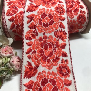 May include: A roll of floral embroidered ribbon. The ribbon features a repeating pattern of large, stylized red flowers with detailed petals and leaves against a white background. The edges are finished with a red border.
