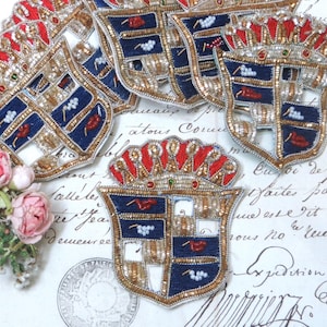 May include: A collection of shield-shaped embroidered patches featuring a blue and gold crest design with red accents, a crown-like top, and small white and red bead details. The patches are arranged in a group with visible metallic gold thread.