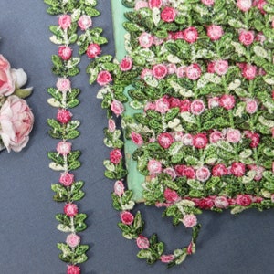 May include: Embroidered floral trim featuring pink roses and green leaves. The trim is arranged in a repeating pattern, with a roll of the trim visible. The trim is suitable for sewing, crafting, and embellishing various projects.
