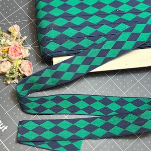 May include: A roll of woven ribbon featuring a repeating diamond pattern in dark navy blue and vibrant green. The ribbon is wrapped around a rectangular cardboard spool, with a loose end extending outward to display the geometric design clearly.
