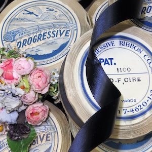 May include: Close-up of spools of white ribbon with blue and black text. The spools are labeled "Progressive Ribbon Co." and "Pat. 1100 D.F. Cire" with a yardage of "7-4620-1-3". A black satin ribbon is wrapped around the spools.
