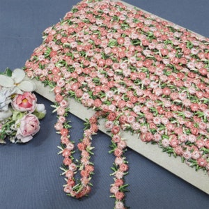May include: A roll of floral trim featuring small, detailed pink and green flowers. The trim is arranged in a dense pattern, with a portion unrolled to show the individual flower strands. The background is a neutral color.
