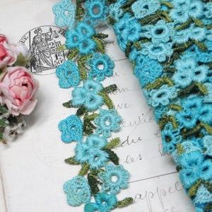May include: A close-up of turquoise blue and green floral lace trim. The lace is delicate and intricate, with small flowers and leaves.