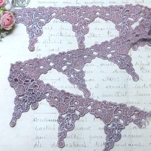 May include: Close-up of mauve lace trim with a scalloped edge. The lace has a delicate floral pattern and a subtle sheen. The trim is ideal for embellishing garments or craft projects. The background is a vintage document.