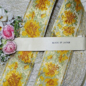 May include: Close-up of a decorative ribbon with a floral pattern in yellow, gold, and green. The ribbon is labeled "MADE IN JAPAN" and is paired with pink and white flowers.