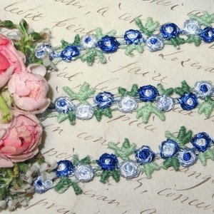 May include: Close-up of decorative floral ribbon featuring blue and white rosettes with green leaves. The ribbon is arranged in rows, with a cluster of pink roses on the left side. The background has faded script.