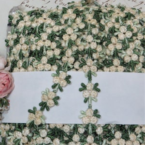 May include: Cream and green floral embroidered trim. The trim features small, clustered cream-colored flowers with green leaves and stems. The trim is densely packed with the floral pattern, creating a textured look. The trim is likely used for embellishing clothing or craft projects.