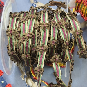 May include: Close-up of a spool of decorative ribbon. The ribbon is brown with vertical stripes of green and purple. Small, decorative brown bows are attached along the edges. The ribbon is in a clear plastic container.