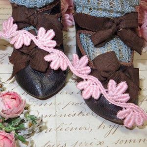 May include: A pair of antique doll shoes with brown leather uppers, adorned with brown ribbon bows and light blue lace. A pink floral lace trim accents the shoes. A small bouquet of pink roses is in the lower left corner.