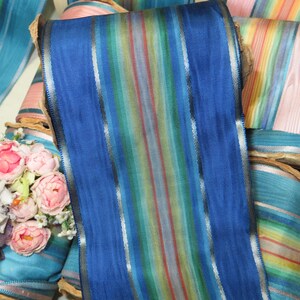 May include: A roll of blue ribbon with vertical stripes in various colors, including red, yellow, and green. The ribbon has a shiny, textured surface and is likely made of silk or a similar material. Other rolls of ribbon in shades of blue and pink are visible.