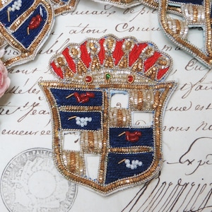 Puede incluir: Un escudo bordado en oro y azul con una corona en la parte superior. El escudo está hecho de cuentas y lentejuelas y tiene un esquema de colores rojo, blanco y azul.