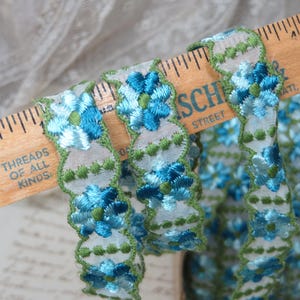 May include: Blue and green floral embroidered trim on a white background. The trim is measured on a wooden ruler with the text "Threads of all kinds".