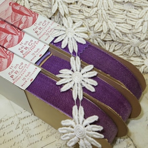 May include: Close-up of vintage purple ribbon spools with white daisy lace trim. The ribbon is labeled "P.R. Co" and "Woven Fast Edge, 10 Yards, Made in France". The background includes more white daisy lace.