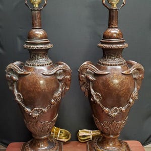 May include: A pair of matching brown table lamps with a faux marble finish. Each lamp features a central urn-shaped body adorned with sculpted ram heads on the sides, floral garland detailing, and a square base. A brass socket sits at the top.