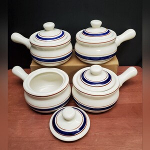 May include: Set of white ceramic soup bowls with lids and handles. Each bowl features a blue and red stripe design around the body and lid. The bowls are arranged on a brown surface, with one lid removed.