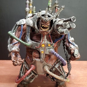 May include: A brown, muscular, monstrous figurine with a metallic chest plate, shoulder-mounted cannon, and various colored tubes connected to its body. The creature features a snarling face with bared teeth and multiple silver exhaust pipes.
