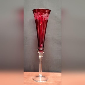 May include: A tall, ruby red champagne flute with a clear stem and base. The top of the glass features a delicate, etched leaf design. The glass has a bulbous section where the stem meets the bowl.