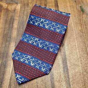 May include: A patterned necktie with a repeating design of red squares and blue stripes. The blue stripes feature a white, stylized design. The tie is made of a smooth fabric and is displayed on a wooden surface.