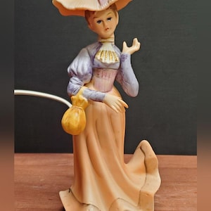 May include: A porcelain figurine of a woman in a long peach dress and a wide-brimmed hat. The figure has a lavender blouse, a pink corset, and a yellow purse. The hat is peach with a pink bow.