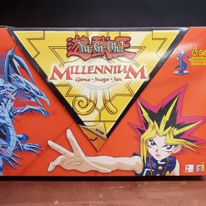 May include: A Yu-Gi-Oh! Millennium Game box with a red background. The box features illustrations of characters and a dragon. Text includes "Yu-Gi-Oh! Millennium Game - Juego - Jeu" and "Action". A small figurine is visible on the box.