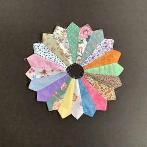 May include: A colourful patchwork quilt block in the shape of a flower with 16 petals. The petals are made of different fabrics with floral patterns in shades of pink, blue, green, and yellow.