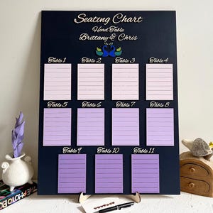 May include: A navy blue seating chart with the text "Seating Chart" and "Head Table Brittany & Chris" in white script. It features 11 rectangular cards in shades of pink and purple, labeled "Table 1" to "Table 11". Two peacock illustrations are in the center.