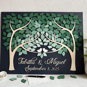 May include: A wooden guest book in the shape of two trees with green leaves. The trees are joined at the top by two white birds. The text "Talitha & Miguel September 8, 2025" is written below the trees.