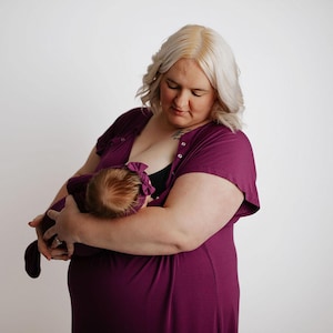 May include: A woman wearing a dark purple nursing dress holds a baby. The baby is wearing a matching purple bow and outfit. The woman has blonde hair and is looking down at the baby.