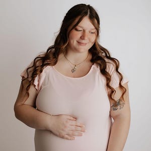 May include: A light pink maternity gown with short, ruffled sleeves. The gown has a scoop neckline and a button closure. A gold necklace with a heart pendant is visible. The person has long, wavy brown hair.