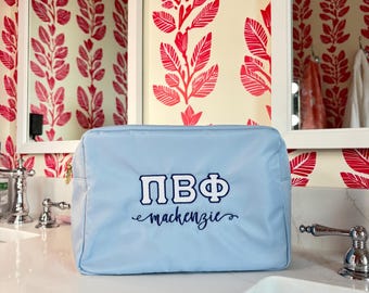 Puede incluir: Un neceser rectangular azul claro con cierre de cremallera. La bolsa presenta las letras griegas ΠΒΦ en blanco con un contorno azul marino, y el nombre "mackenzie" en escritura azul marino. La bolsa está sobre una superficie blanca.