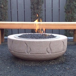 May include: A stone fire pit with a carved design and a small fire burning inside. The fire pit is sitting on a bed of gravel. A wooden bench is in the background.