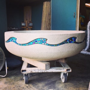 May include: A large, white concrete planter with a mosaic wave design in shades of blue and turquoise. The planter is sitting on a metal dolly with wheels.