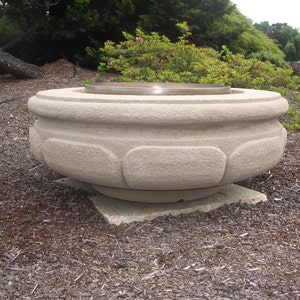 May include: A large, round, light-colored stone planter with a smooth, textured surface. The planter has a wide, flat base and a slightly flared top. The planter is sitting on a bed of brown mulch.