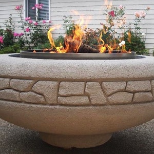 May include: A large, round, concrete fire pit with a stone-like texture. The fire pit has a metal ring around the top and a fire burning inside.